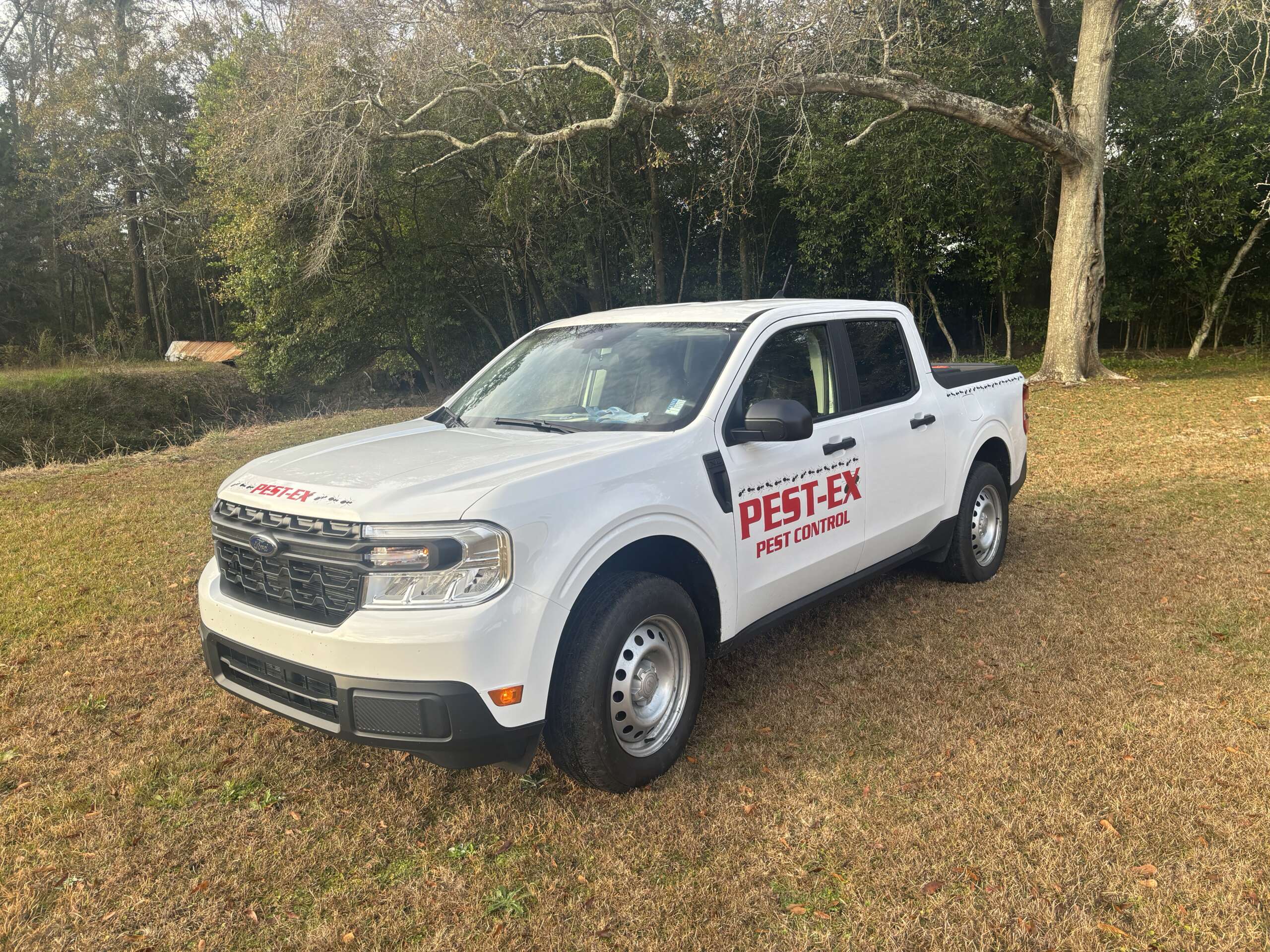 A white pickup truck with PEST-EX Pest Control written on the door and hood is parked on grass in a rural area with trees in the background.