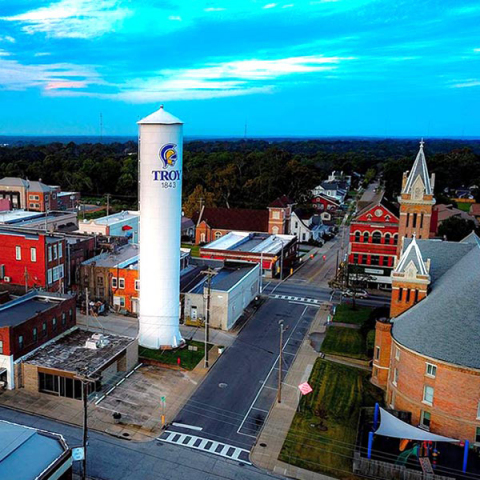 Aerial view of a small town with a tall white water tower labeled Troy 1843, surrounded by buildings, streets, and a church with a clock tower, under a blue sky.